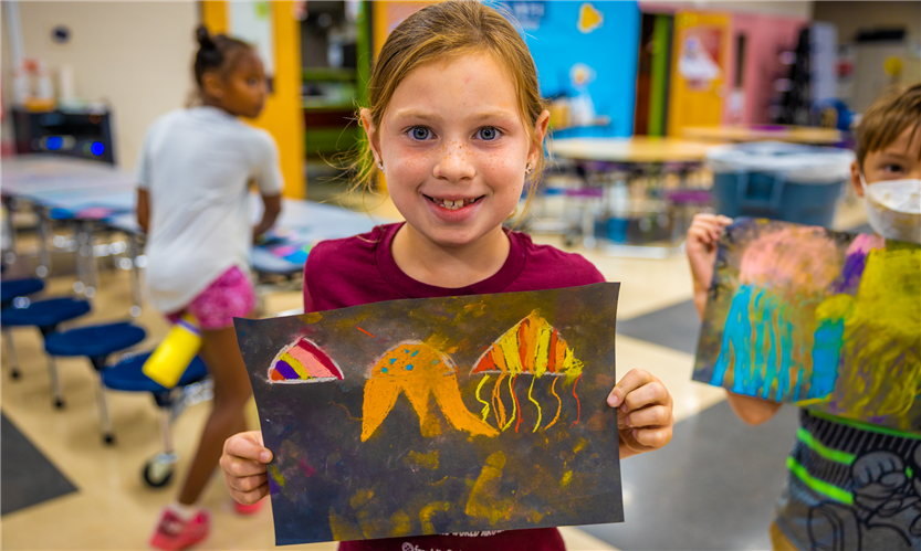 A girl wearing a red shirt holds up a piece of art